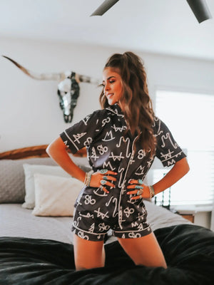 Full-length view of women's black pajama set with shorts, showing silky fabric and white brand print in Western bedroom setting